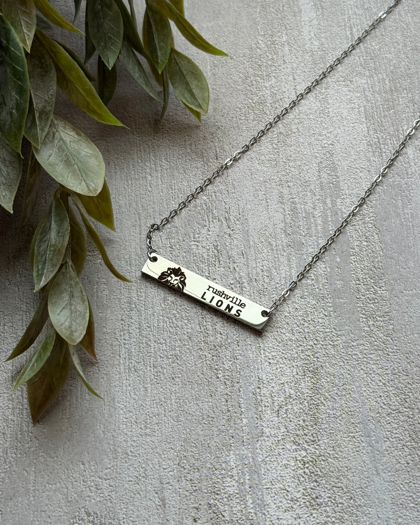 Silver necklace with a bar pendant on a light gray surface with green leaves.