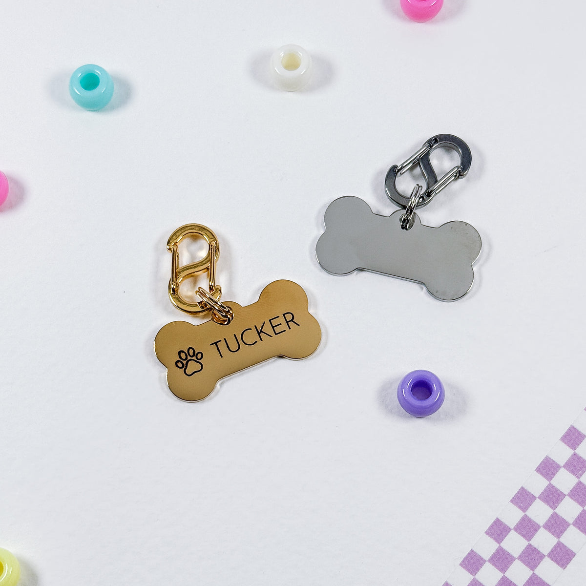 Two bone-shaped pet ID tags on a white background, one gold with the name 'TUCKER' and paw print, and one silver. Small colorful beads are scattered around as props.