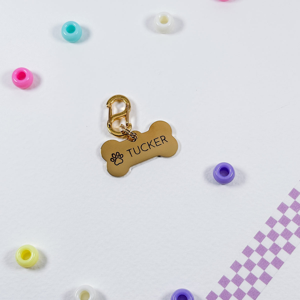 A gold-colored bone-shaped pet ID tag engraved with the name 'TUCKER' and a paw print, displayed on a white background with colorful beads and a purple checkered strip.