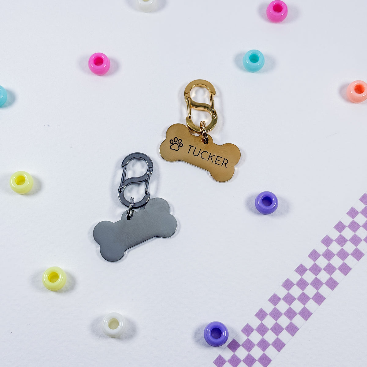 Two personalized pet ID tags shaped like bones, one gold and one silver, with carabiner clips. One tag is engraved with a paw print and the name 'TUCKER'.