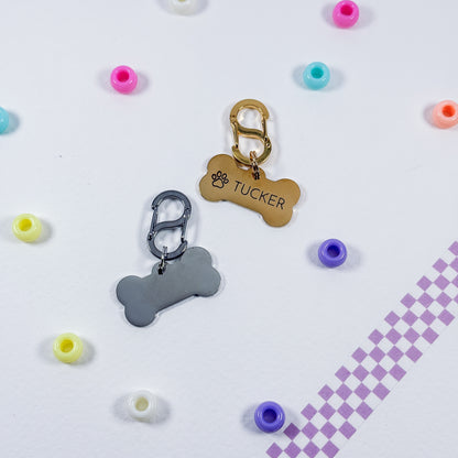 Two personalized pet ID tags shaped like bones, one gold and one silver, with carabiner clips. One tag is engraved with a paw print and the name 'TUCKER'.