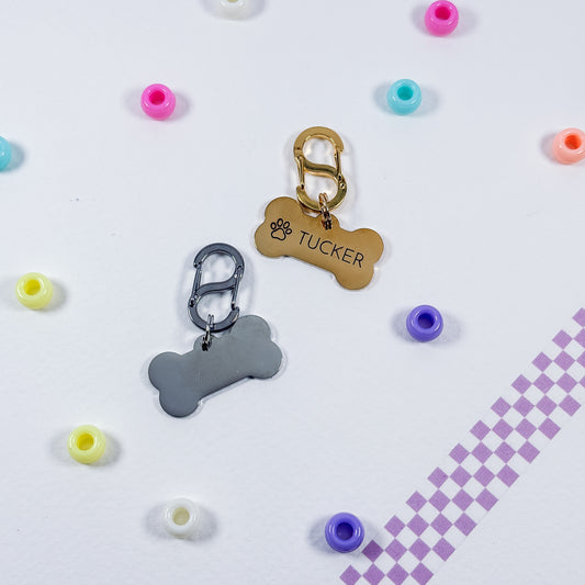 Two personalized pet ID tags shaped like bones, one gold and one silver, with carabiner clips. One tag is engraved with a paw print and the name 'TUCKER'.