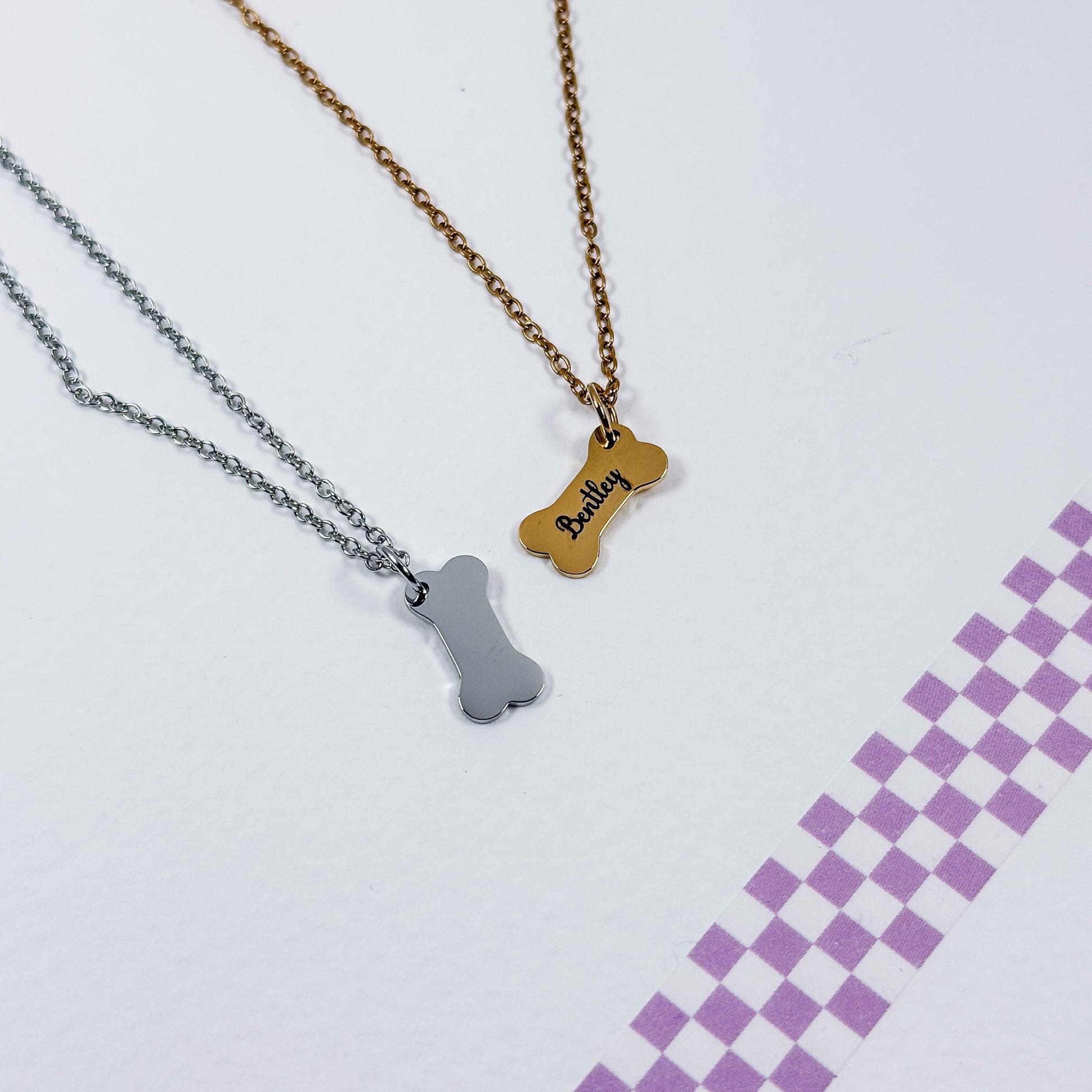 Two dog bone-shaped necklaces, one gold and one silver, on a light purple background with a checkered pattern.