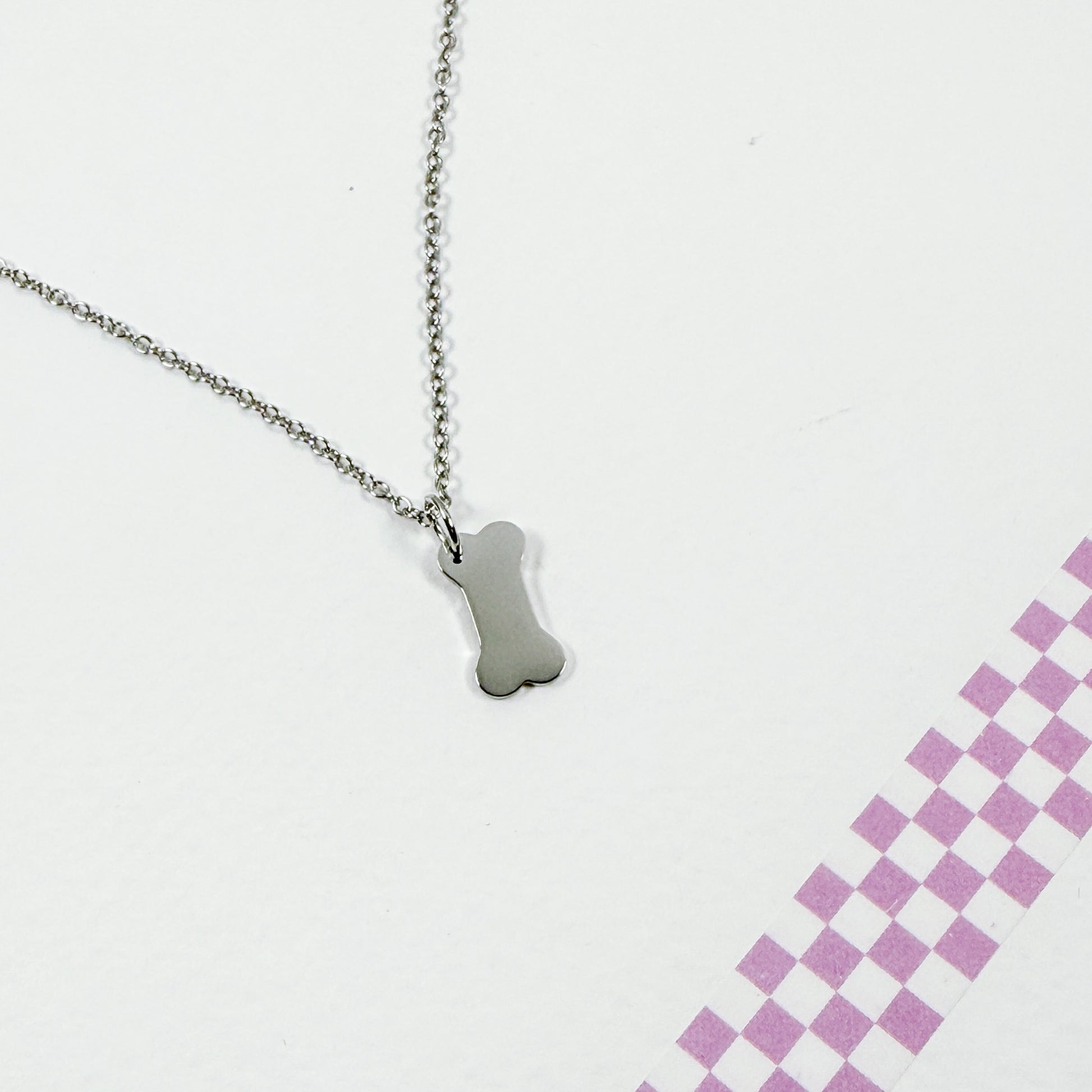 Silver bone-shaped pendant on a chain against a white background with a pink checkered pattern.