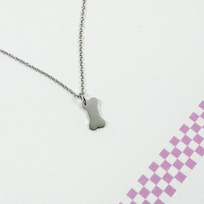 Silver bone-shaped pendant on a chain against a white background with a pink checkered pattern.