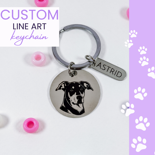 A silver metal keychain with a round engraved disc featuring a black line art dog portrait and an elongated tag engraved with the name ASTRID. The keychain is displayed on a white surface with pink beads, alongside a purple sidebar with white paw prints and a text overlay reading 'CUSTOM LINE ART keychain'.