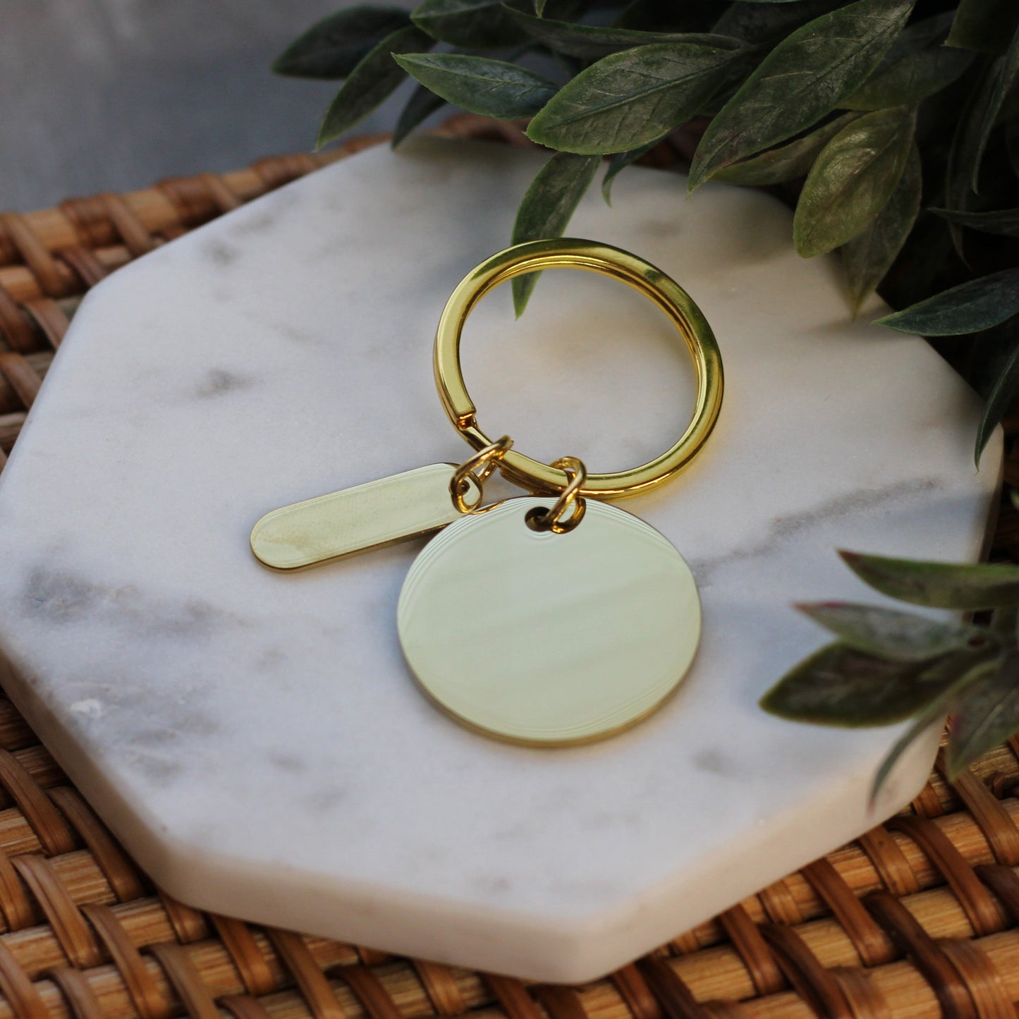 Gold keychain with two round tags on a marble coaster with green leaves in the background