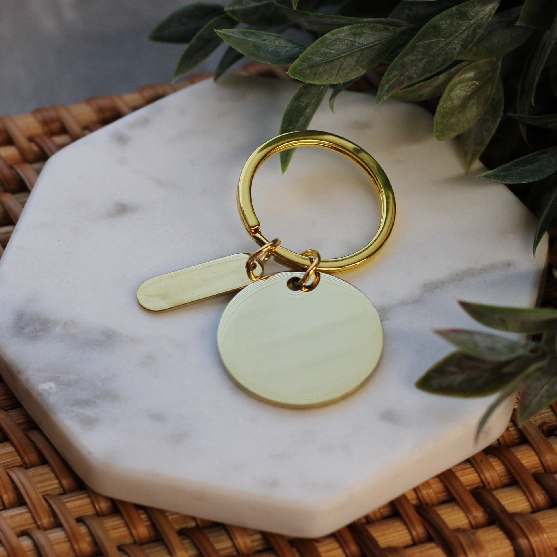 Gold keychain with two round tags on a marble coaster with green leaves in the background