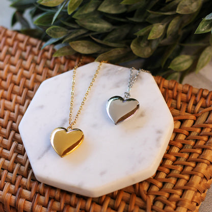 Gold and silver heart-shaped pendants on a marble stand with green leaves in the background.