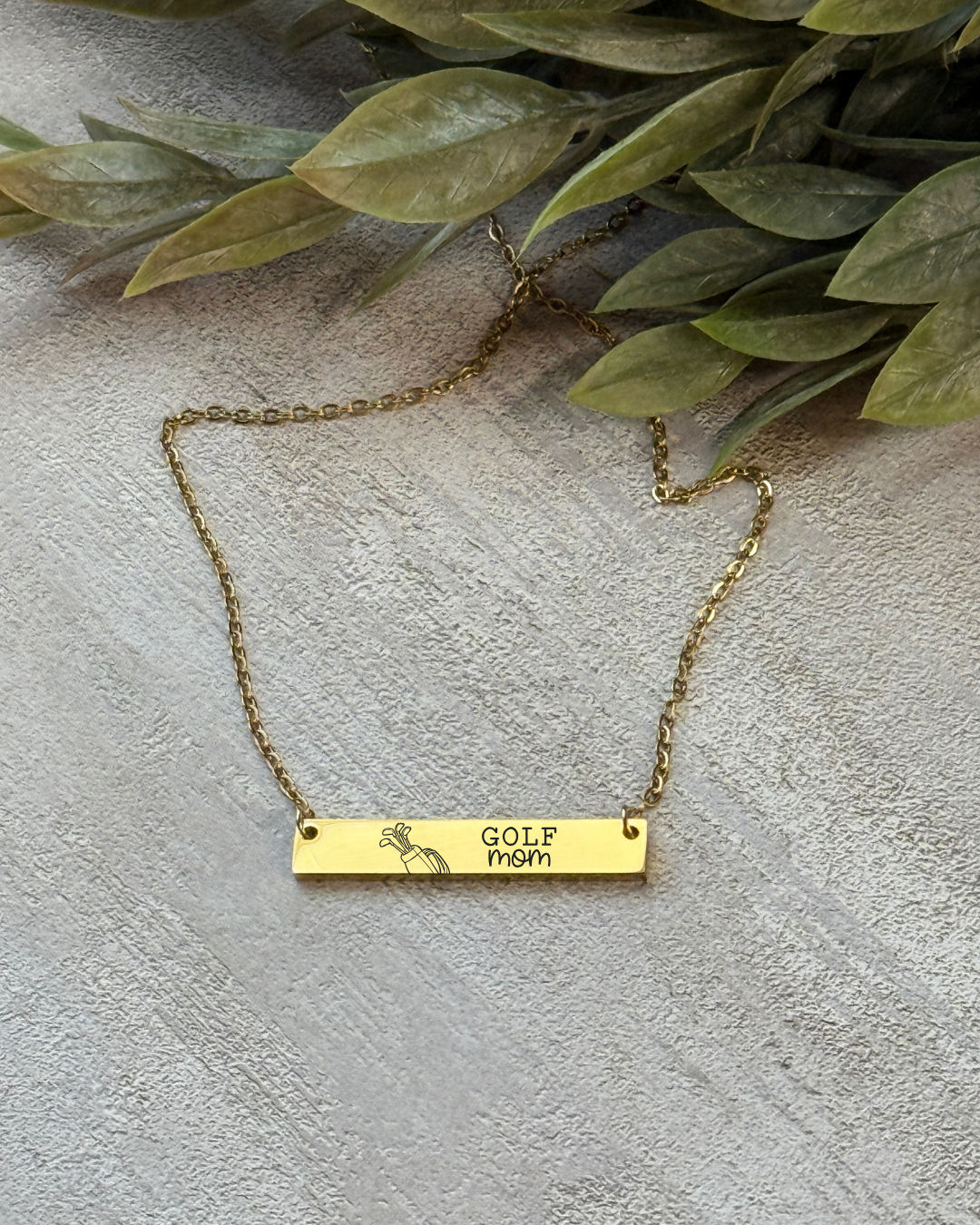 Gold necklace with a bar pendant on a textured surface with green leaves.