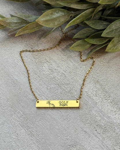 Gold necklace with a bar pendant on a textured surface with green leaves.