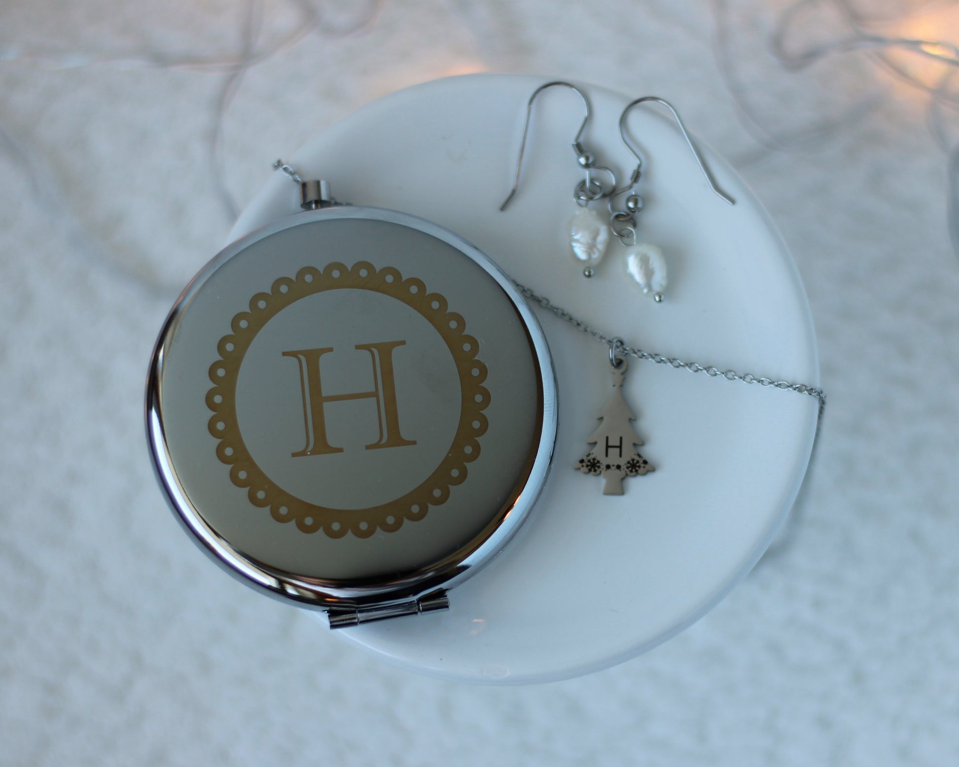 Jewelry set including a compact mirror with a 'H' emblem, necklace, and earrings on a textured white surface.