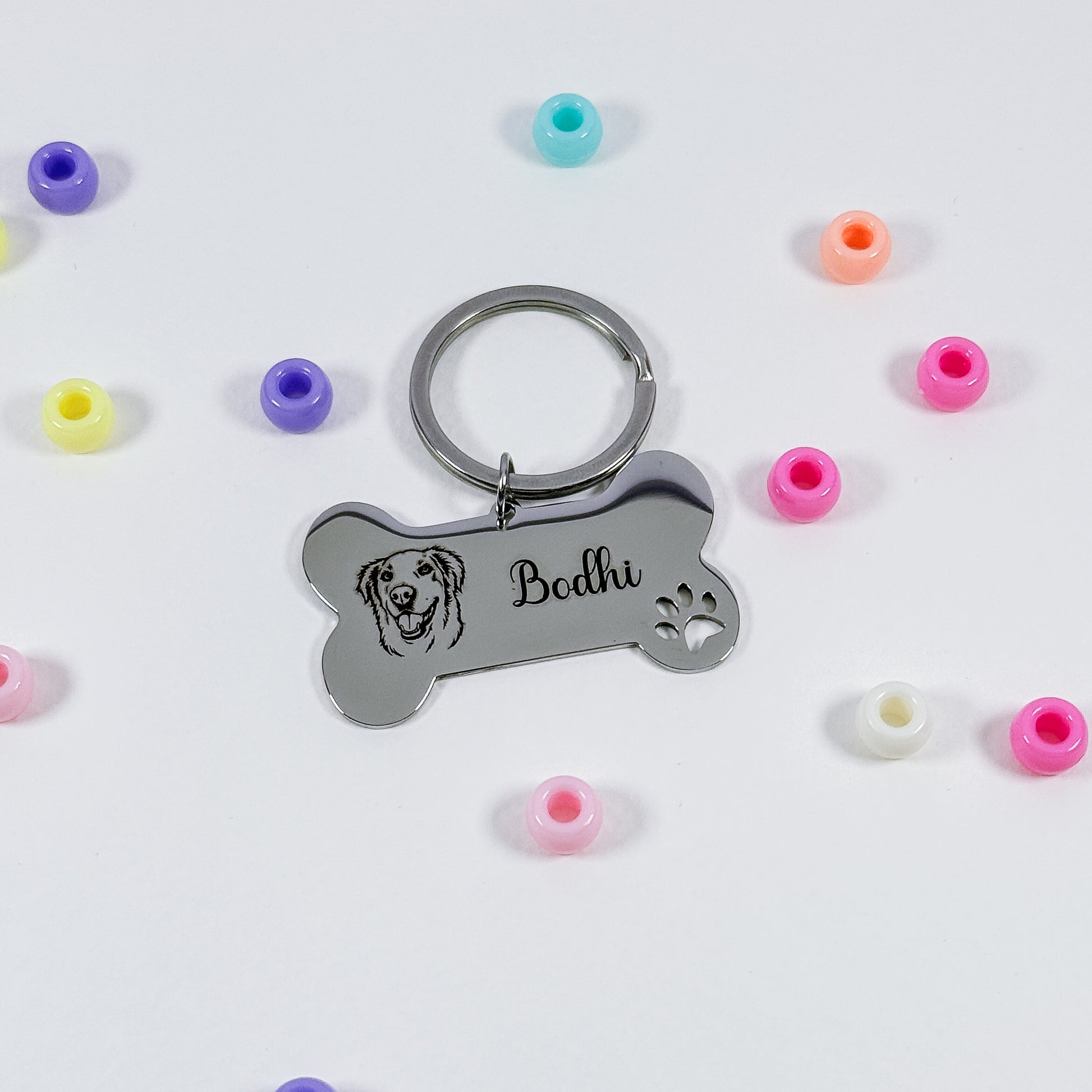 A silver metal bone-shaped keychain with an engraved dog face, the name 'Bodhi', and a paw print, shown on a white background with scattered colorful silicone beads.