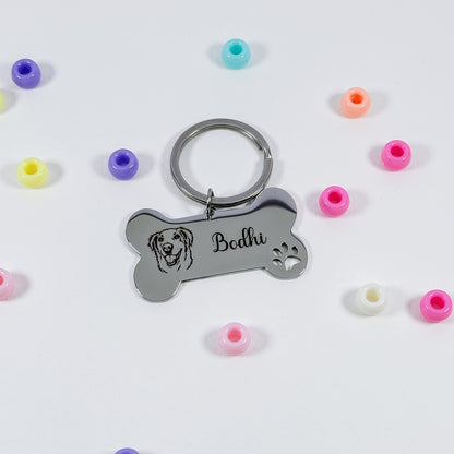 A silver metal bone-shaped keychain with an engraved dog face, the name 'Bodhi', and a paw print, shown on a white background with scattered colorful silicone beads.
