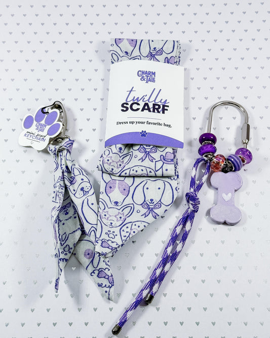 A pet accessory set featuring a fabric pet scarf with a purple and white cartoon dog pattern, shown both folded in its packaging and hanging from a silver carabiner with purple beads and a bone-shaped charm. The brand 'Chic & Star Pet Supplies' is visible on the packaging and tag.