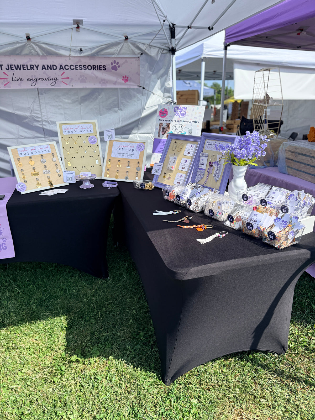 An outdoor market booth under a white canopy tent displaying jewelry and accessories. The black-covered tables hold framed product cards, wrapped items, and a vase of purple flowers on the grass.