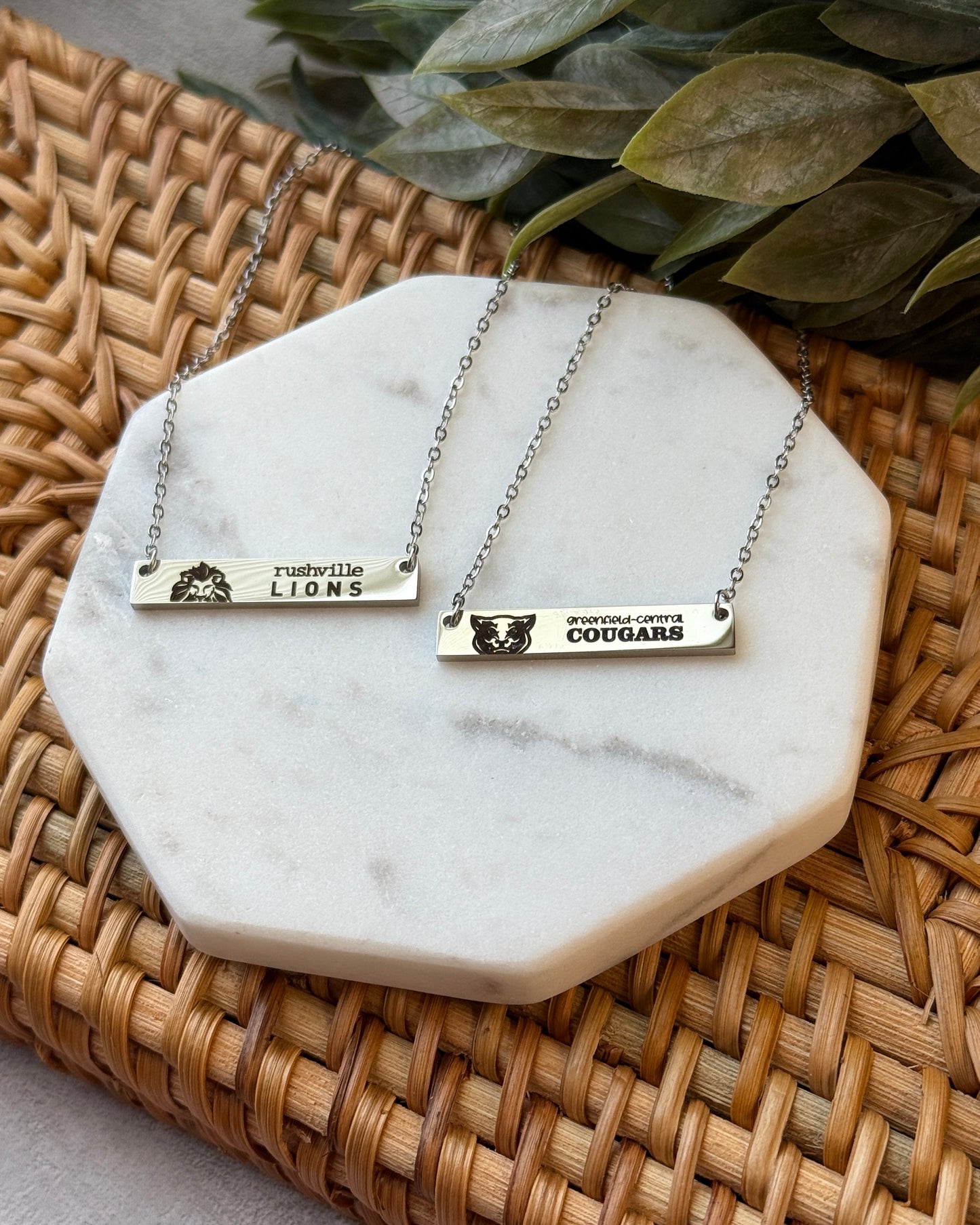 Two necklaces with engraved names on a marble coaster with a woven surface and green leaves in the background.