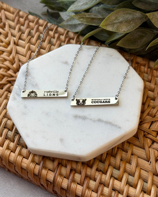 Two necklaces with engraved names on a marble coaster with a woven surface and green leaves in the background.