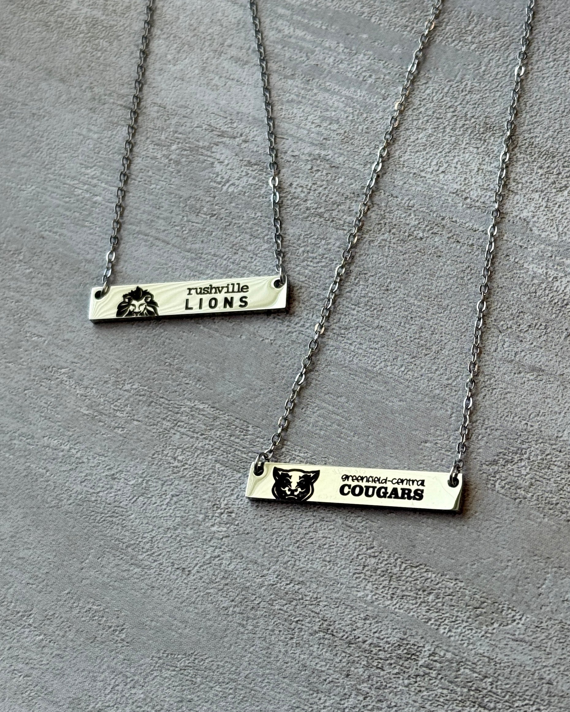 Two silver bar necklaces with engraved text on a gray surface