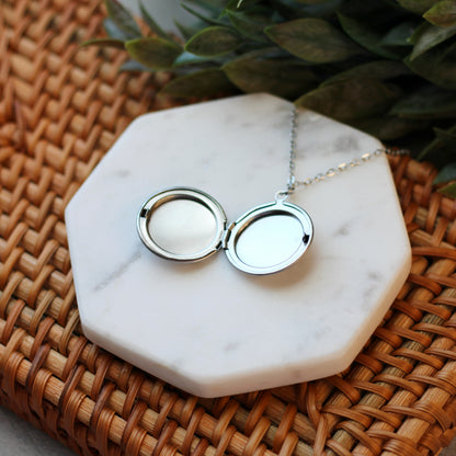 Silver locket necklace on a marble stand with a woven surface and green leaves in the background
