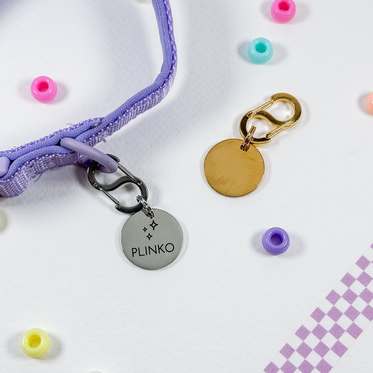 A close-up flat lay of a lavender purple woven strap with a silver round metal charm engraved with the name 'PLINKO' and a gold round disc charm with a clasp, surrounded by colorful beads on a white background.