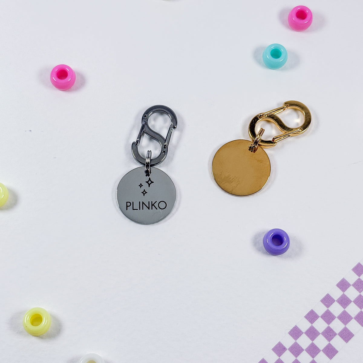 Two round metal keychains with carabiners are displayed on a white surface with colorful beads scattered around. One keychain has a silver-toned disc engraved with the name 'PLINKO' and a star motif, and the other has a blank gold-toned disc.
