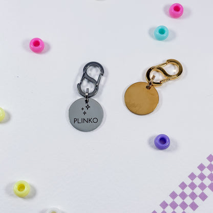 Two round metal keychains with carabiners are displayed on a white surface with colorful beads scattered around. One keychain has a silver-toned disc engraved with the name 'PLINKO' and a star motif, and the other has a blank gold-toned disc.
