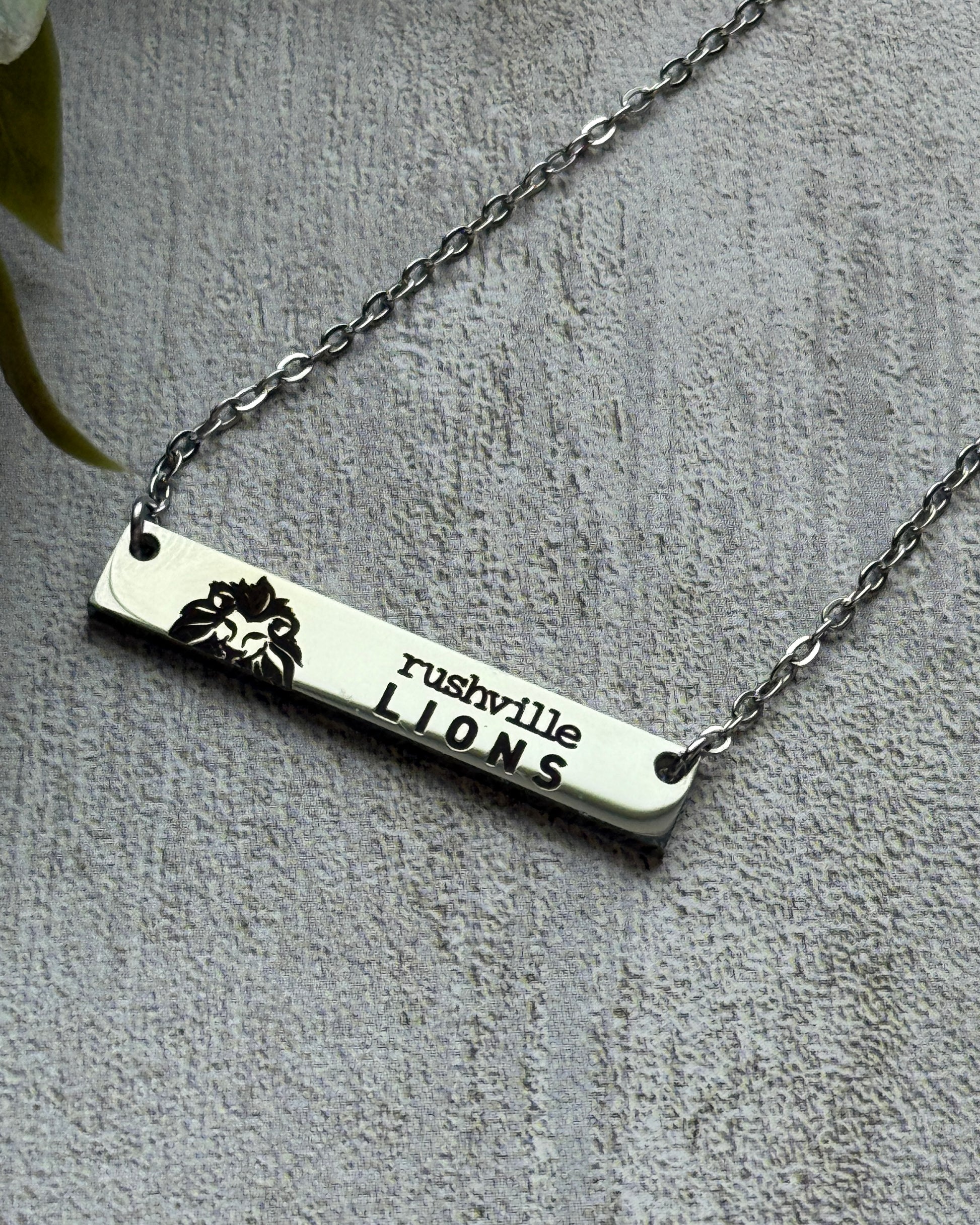 Silver necklace with 'rushville LIONS' bar pendant on a gray background