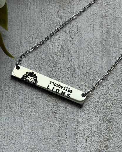 Silver necklace with 'rushville LIONS' bar pendant on a gray background