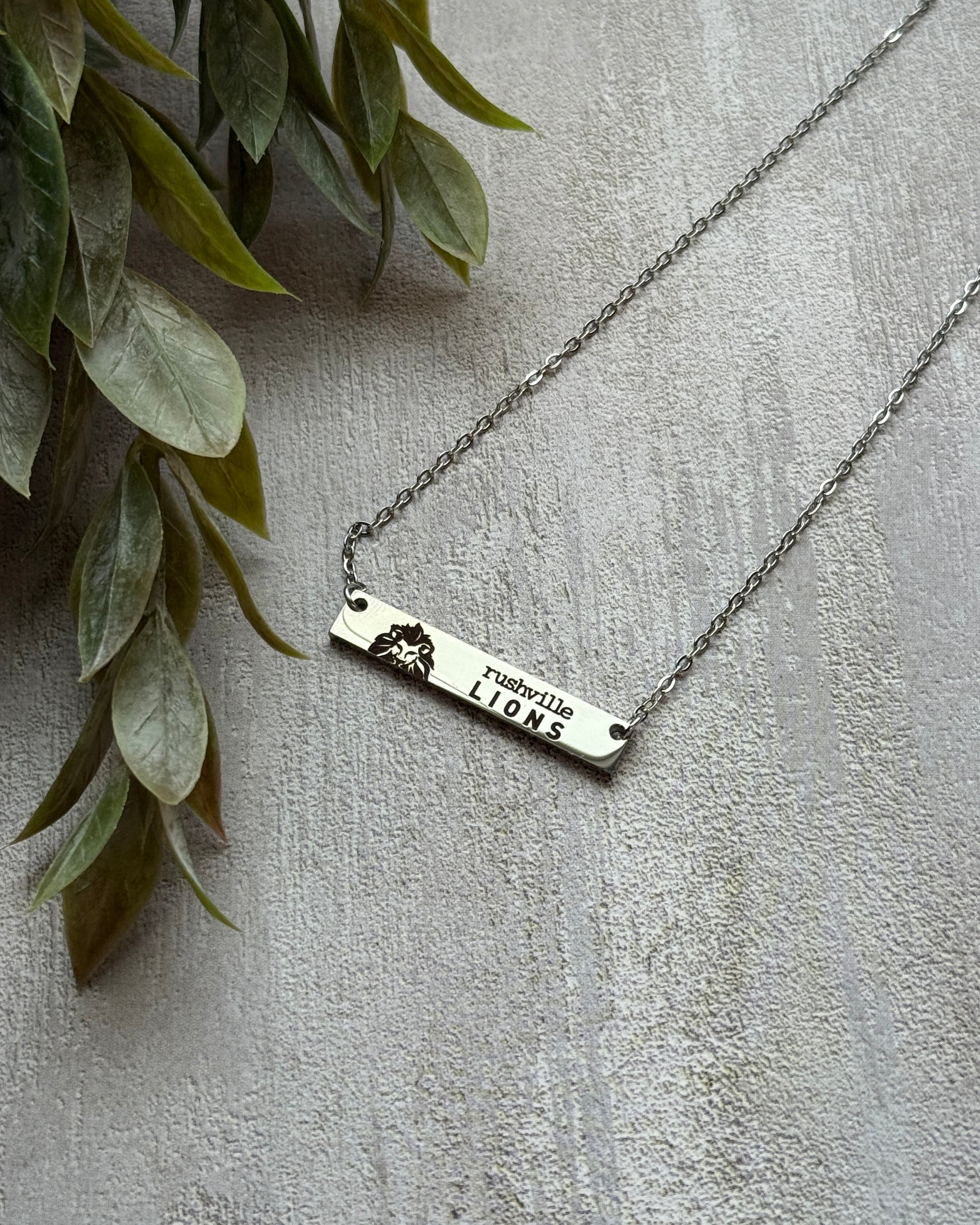 Silver necklace with a bar pendant on a light gray surface with green leaves.