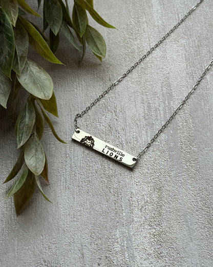 Silver necklace with a bar pendant on a light gray surface with green leaves.