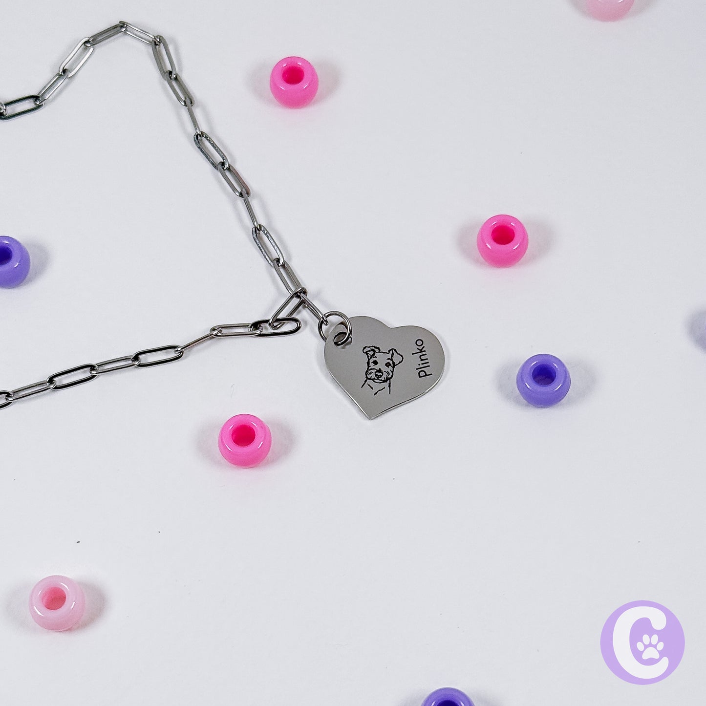 Bracelet with heart-shaped charm featuring a dog illustration and 'Plinko' text, surrounded by colorful beads on a light background.