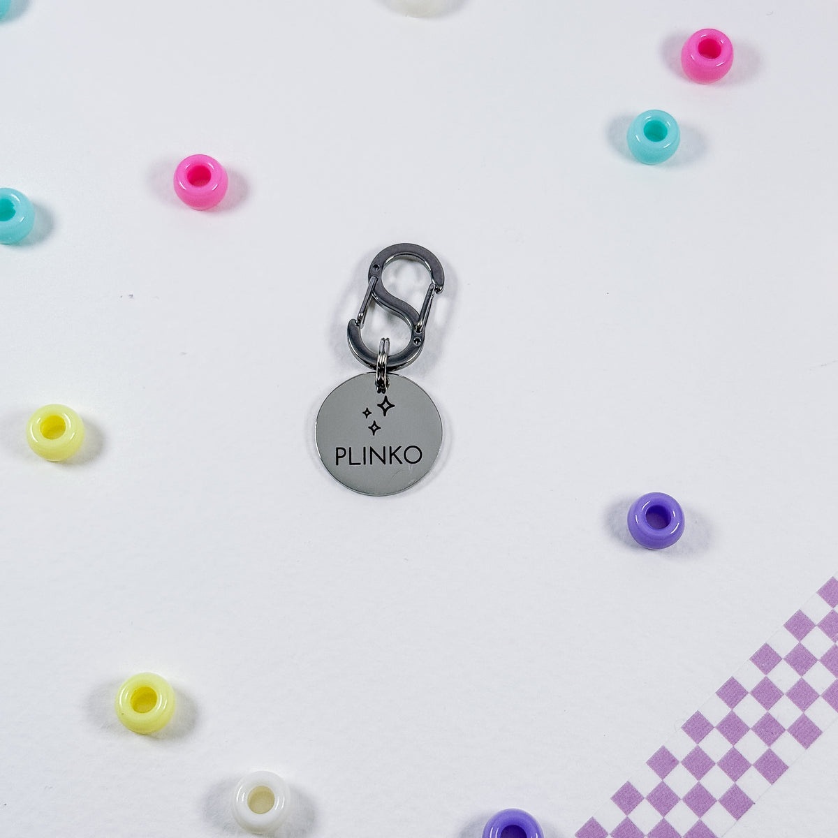 A silver metal charm keychain with a round disc engraved with the name 'PLINKO' and small star details, attached to a dark gray carabiner. The keychain is displayed on a white surface surrounded by scattered pastel-colored beads, with a purple checkered pattern in the bottom right corner.