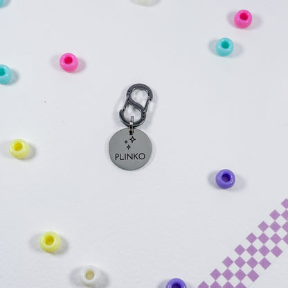 A silver metal charm keychain with a round disc engraved with the name 'PLINKO' and small star details, attached to a dark gray carabiner. The keychain is displayed on a white surface surrounded by scattered pastel-colored beads, with a purple checkered pattern in the bottom right corner.