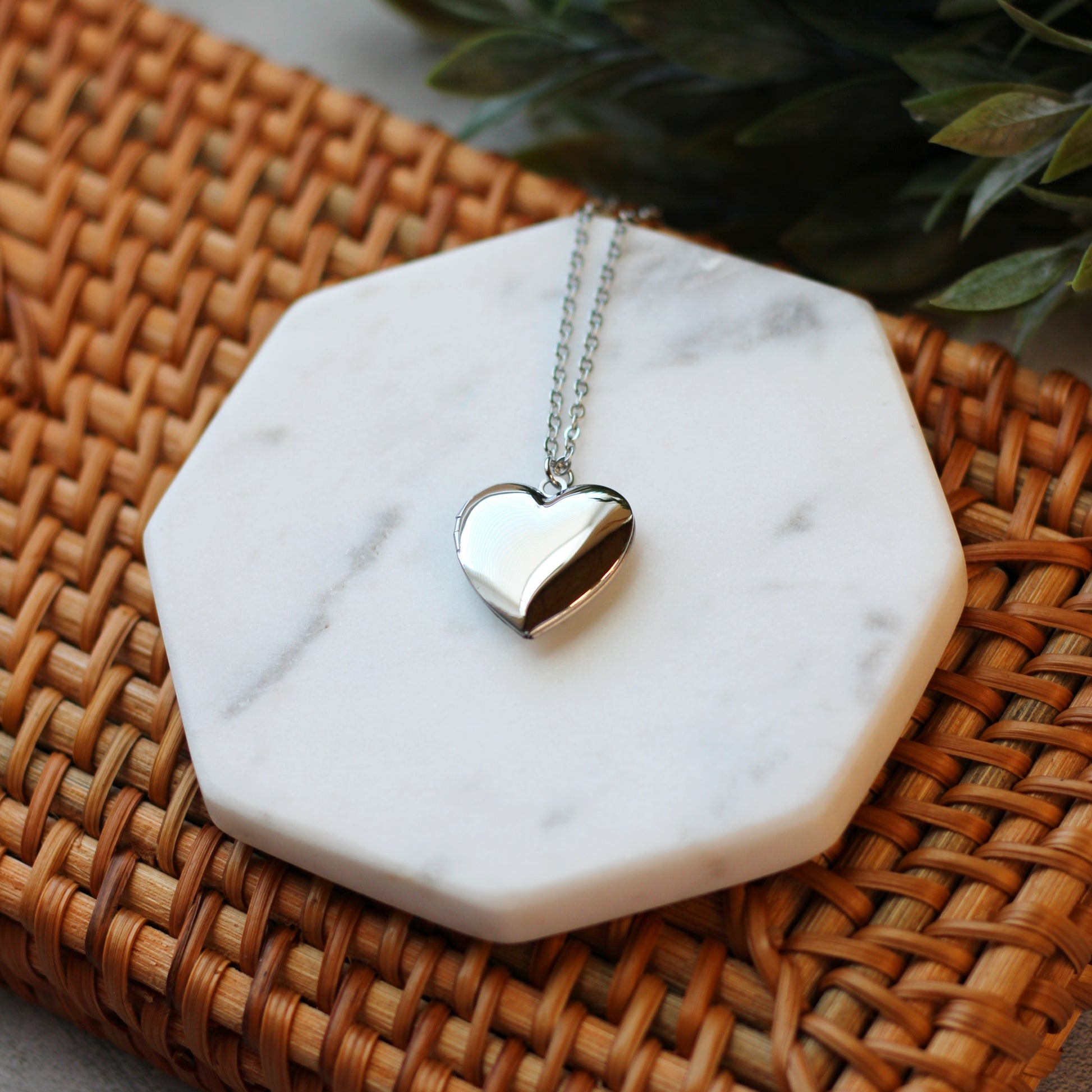 Silver heart-shaped pendant necklace on a marble stand with a woven surface and green leaves in the background