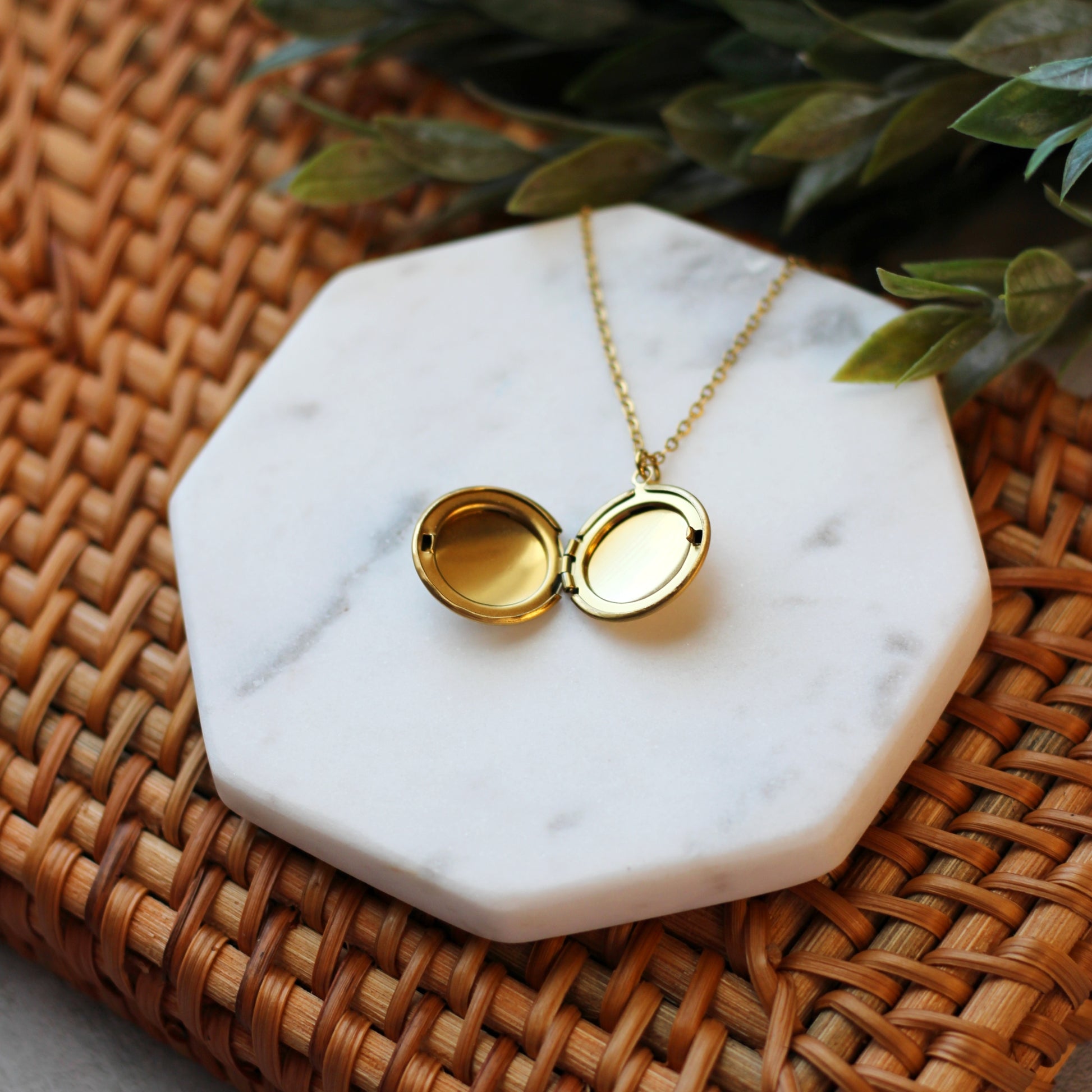 Gold locket necklace on a marble coaster with a woven surface and green leaves in the background