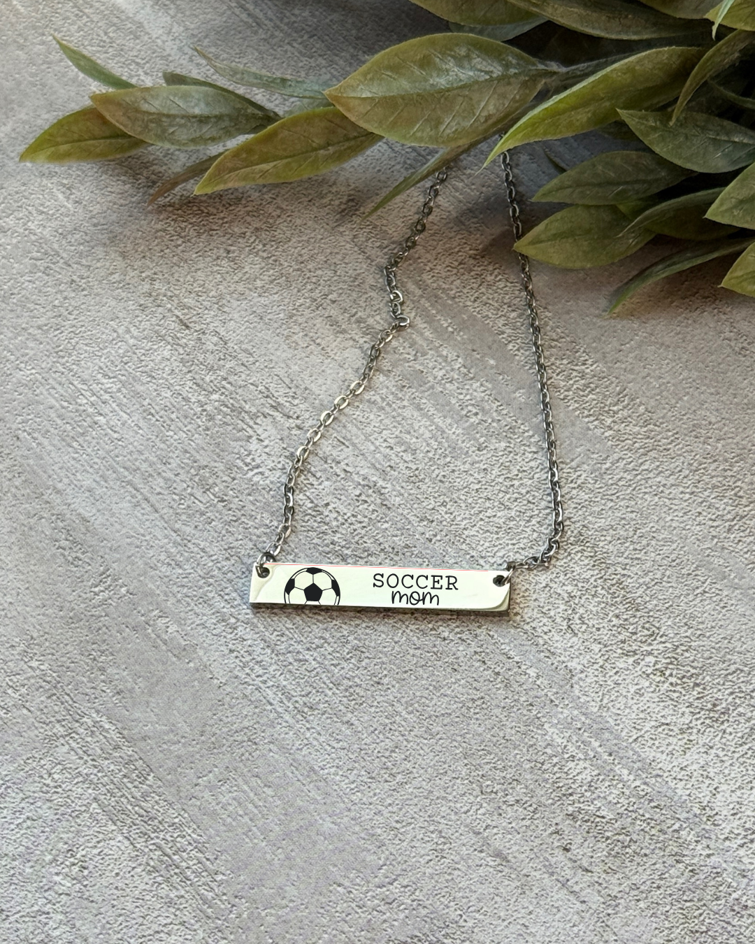 Necklace with 'Soccer Mom' bar pendant on a textured surface with green leaves.