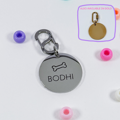 A round silver metal pet ID tag with a bone icon and the name 'BODHI' engraved, attached to a silver clasp. The image includes small colorful beads around it and a purple inset showing the tag in gold.
